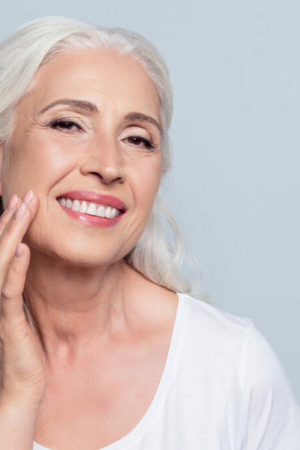 Charming, pretty, old woman touching her perfect soft face skin with fingers, smiling at camera over gray background, using day, night face cream, cosmetology procedures